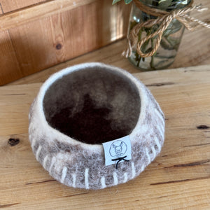 Felted bowl with a brand tag on a wooden surface, next to a vase with greenery.