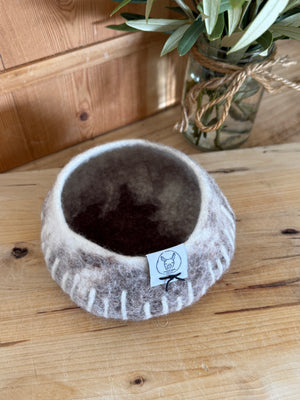 Felted bowl with a brand tag on a wooden surface, next to a vase with greenery.