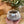 Hand holding a textured felted bowl with a label, placed on a wooden surface with plants in the background.