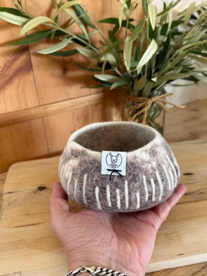 Hand holding a textured felted bowl with a label, placed on a wooden surface with plants in the background.